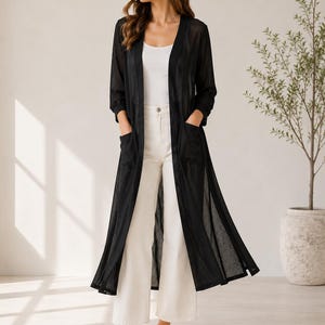 May include: A long, sheer black duster cardigan featuring three-quarter length sleeves and two front patch pockets, worn over a white tank top and white wide-leg trousers, paired with simple tan slide sandals for a layered ensemble.