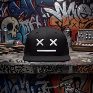May include: A black baseball cap featuring a white embroidered graphic on the front panel. The design consists of two white crosses positioned as eyes and a single horizontal white line underneath, representing a neutral or deadpan facial expression.
