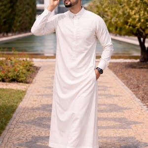 Op de afbeelding: Een witte, lange mouwen thobe, een traditioneel Arabisch kledingstuk, gedragen met witte sneakers. De thobe is enkellang en heeft een button-down kraag. De persoon draagt een zonnebril en een zwart polshorloge. De achtergrond omvat bomen en een gebouw.