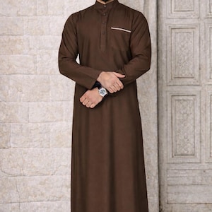 May include: A long, brown thobe with a mandarin collar and a small pocket detail. The traditional garment features long sleeves and a button-down front. The model is wearing a watch and black shoes. The background is a light-colored brick wall and a wooden door.