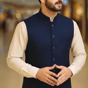 Op de afbeelding: Een marineblauwe vest met een opstaande kraag en knoopsluiting, gedragen over een crèmekleurig shirt met lange mouwen. De outfit wordt gecompleteerd met een bijpassende crèmekleurige broek, wat een formeel ensemble creëert.