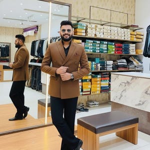 Pode incluir: Um homem com um blazer marrom de abotoamento duplo, cal&ccedil;as pretas e sapatos pretos. O blazer tem um len&ccedil;o de bolso. O fundo mostra uma loja de roupas com prateleiras de camisas dobradas e caixas.