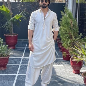 May include: A cream-colored, long-sleeved tunic and matching loose-fitting pants. The outfit is paired with black sandals. The person is wearing sunglasses and a watch. The background includes potted plants and a tiled patio.