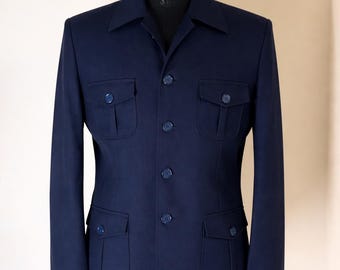 May include: A dark navy blue jacket featuring a classic collar, a central button-down closure, and four front patch pockets with buttoned flaps. The long sleeves are finished with small buttons at the cuffs, and the garment is displayed on a mannequin.