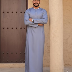 Può includere: Un thobe azzurro, un tradizionale indumento arabo, con finiture bianche sul colletto e sui polsini. Il thobe è indossato con sandali marroni. La persona è in piedi davanti a una porta marrone e una colonna beige.