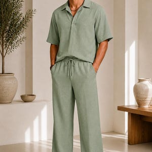 May include: A sage green short-sleeved shirt with a collar and a partial button placket, paired with matching sage green wide-leg trousers featuring an elasticized waistband and a drawstring closure, worn with brown leather slide sandals.