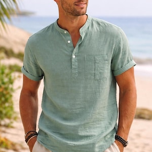 May include: A sage green short-sleeved shirt with a mandarin collar, a partial button-down front placket, and a single chest pocket. The shirt features rolled-up sleeves and a textured fabric finish, paired with light beige drawstring shorts.