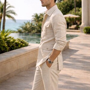May include: A light beige linen suit, including a button-down shirt with two chest pockets and matching pants. A silver wristwatch is visible. The setting is outdoors with palm trees and a pool in the background. The suit appears to be made of a lightweight fabric.
