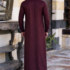Pu&ograve; includere: Una thobe a maniche lunghe che arriva fino ai piedi in un colore bordeaux profondo e uniforme. L'indumento presenta un colletto alto, una silhouette dal taglio dritto e polsini con bottoni. Il tessuto ha una finitura liscia e opaca.