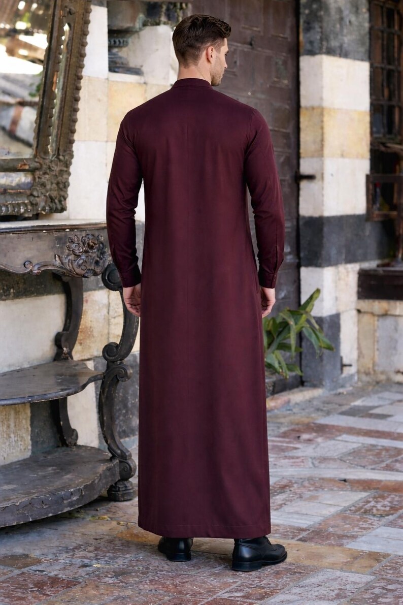 Pu&ograve; includere: Una thobe a maniche lunghe che arriva fino ai piedi in un colore bordeaux profondo e uniforme. L'indumento presenta un colletto alto, una silhouette dal taglio dritto e polsini con bottoni. Il tessuto ha una finitura liscia e opaca.