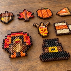 May include: A collection of eight autumn-themed fuse bead designs including an acorn, a leaf, a pumpkin, a slice of pie, a turkey, a corn cob, a mug, and a pilgrim hat, all crafted from small, colorful, circular plastic beads.