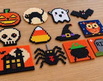 Op de afbeelding: Een verzameling van twaalf Halloween-ontwerpen gemaakt van strijkkralen, waaronder een pompoen, een zwarte kat, een spook, een vleermuis, een schedel, een candy corn, een heksenhoed, een uil, een spookhuis, een spin, een ketel en een grafsteen met de tekst RIP.