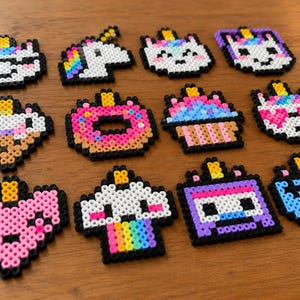 Może przedstawiać: Kolekcja dwunastu kolorowych figurek w stylu pixel art wykonanych z małych plastikowych koralików. Wzory obejmują różne głowy jednorożców, różowego pączka, babeczkę, serce, tęczową chmurę, fioletowy telewizor oraz niebieski kontroler do gier wideo.