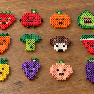 May include: A collection of twelve colorful fruit and vegetable figures crafted from plastic fuse beads. Each piece features a simple, pixelated design with a smiling face, including representations of a strawberry, apple, orange, avocado, carrot, broccoli, mushroom, watermelon, pineapple, grapes, peach, and eggplant.