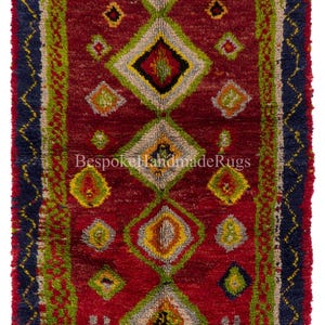 May include: A rectangular, handmade rug with a deep red background and geometric patterns. The design features diamond shapes in yellow, green, and gray, framed by a navy blue border with a zigzag pattern. The text "Bespoke Handmade Rugs" is visible.