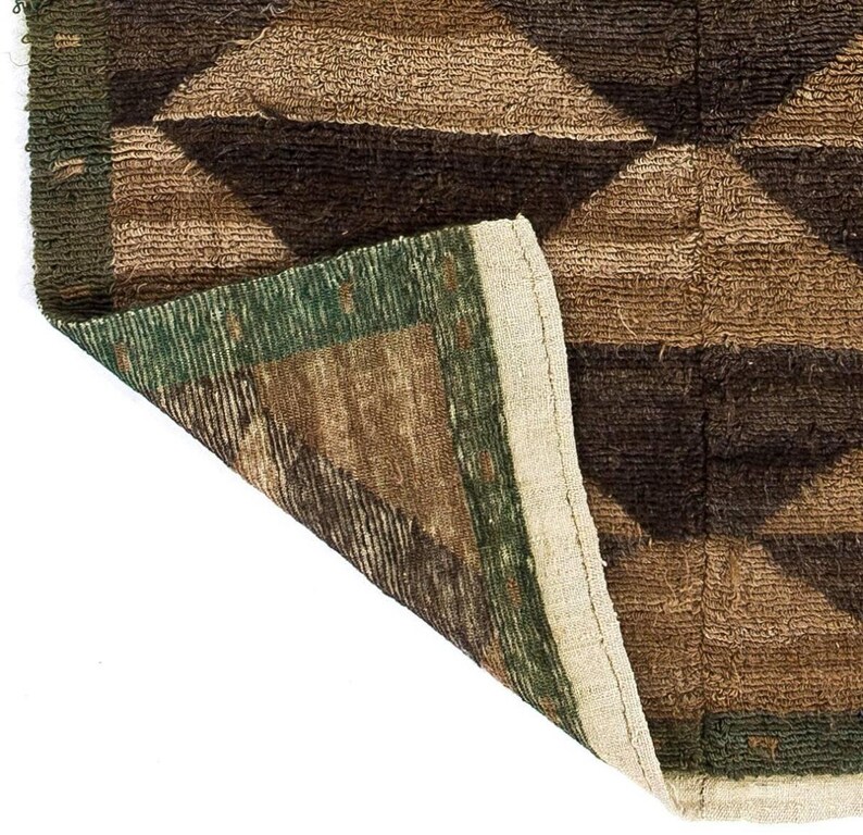May include: Close-up of a brown and green geometric patterned rug. The rug features a diamond pattern in shades of brown and green, with a cream-colored border. The rug's texture appears to be woven.
