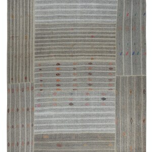 Puede incluir: Una alfombra rectangular gris con un diseño de patchwork. La alfombra presenta rayas verticales y horizontales en diferentes tonos de gris y blanco. Pequeños rombos de colores están dispersos por la superficie, añadiendo un toque de color a la paleta neutra.