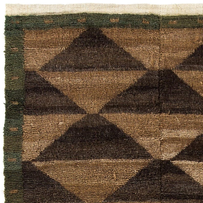 May include: Close-up of a textured rug with a geometric pattern. The rug features alternating brown and dark brown triangles. A green border frames the design, with a cream-colored edge.