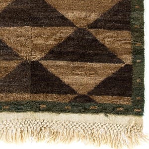 May include: Close-up of a brown and black geometric patterned rug. The rug features a repeating diamond pattern in shades of brown and black. A green border frames the design, and the rug has a fringe along the bottom edge.