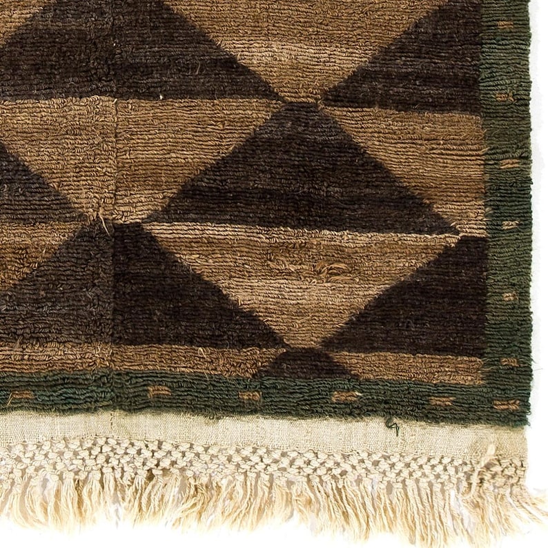 May include: Close-up of a brown and black geometric patterned rug. The rug features a repeating diamond pattern in shades of brown and black. A green border frames the design, and the rug has a fringe along the bottom edge.