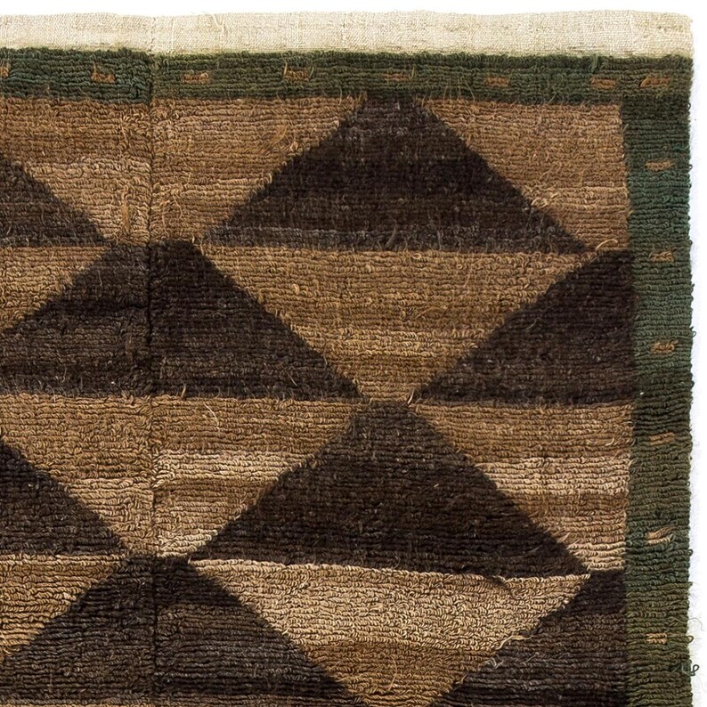 May include: Close-up of a woven textile with a geometric pattern. The design features dark brown and tan triangles arranged in a repeating pattern. A green border frames the design, and the edge has a natural, off-white fringe.