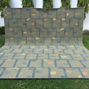 Pode incluir: Um tapete retangular grande com uma base verde-oliva suave e um padrão de grade formado por linhas azul-escuras. O design inclui pequenos motivos geométricos espalhados em tons de laranja, amarelo e azul dentro das seções da grade.