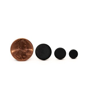May include: Three black round wooden discs of varying sizes, next to a US penny for scale.