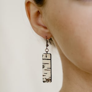 May include: A pair of dangle earrings with a rectangular birch bark design. The earrings are made of copper and have a simple hook closure.