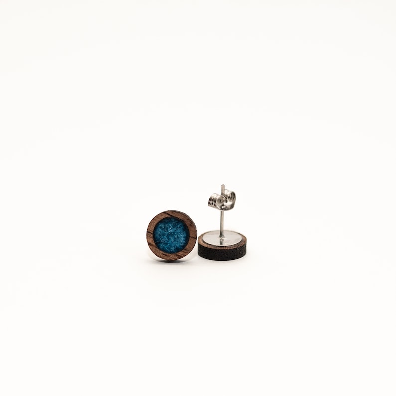 Peut inclure: Une paire de boucles d'oreilles rondes. Chaque boucle d'oreille pr&eacute;sente un cadre en bois fonc&eacute; avec un centre de couleur teal. Les tiges des boucles d'oreilles sont en m&eacute;tal argent&eacute;. Les boucles d'oreilles sont pr&eacute;sent&eacute;es sur fond blanc.