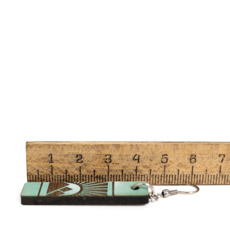 K&ouml;nnte beinhalten: Ein Holzohrring mit einem t&uuml;rkis-braunen geometrischen Design, etwa 5 cm lang. Der Ohrring wird neben einem Holzlineal zur Gr&ouml;&szlig;enbestimmung gezeigt, mit einem silberfarbenen Ohrhaken.