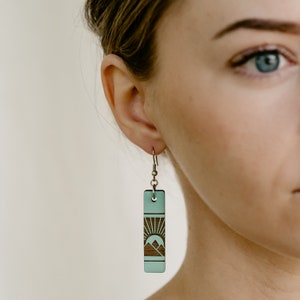 K&ouml;nnte beinhalten: Ein einzelner rechteckiger Ohrring mit einem Berg- und Sonnendesign. Der Ohrring hat eine hellt&uuml;rkise Farbe mit einem dunkelbraunen Berg- und Sonnendesign. Der Ohrring h&auml;ngt an einem silbernen Haken.
