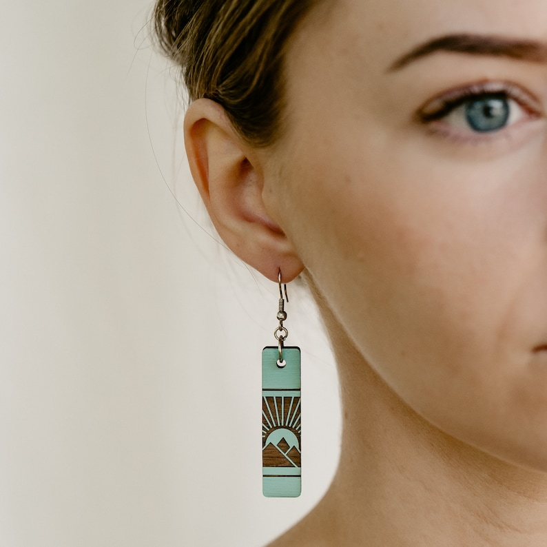 K&ouml;nnte beinhalten: Ein einzelner rechteckiger Ohrring mit einem Berg- und Sonnendesign. Der Ohrring hat eine hellt&uuml;rkise Farbe mit einem dunkelbraunen Berg- und Sonnendesign. Der Ohrring h&auml;ngt an einem silbernen Haken.