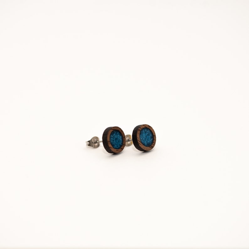 Peut inclure: Deux boucles d'oreilles rondes en bois, avec un cadre marron fonc&eacute; et un centre bleu turquoise. Les boucles d'oreilles ont un design simple et minimaliste, sur fond blanc. Les tiges sont argent&eacute;es.
