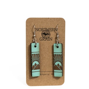 K&ouml;nnte beinhalten: Rechteckige Ohrringe mit einem Berg- und Sonnendesign. Die Ohrringe sind t&uuml;rkis mit einem dunkelbraunen Design und silbernen Haken. Sie werden auf einer braunen Karte mit dem Text "NORTHERN GRAIN" und einem Logo pr&auml;sentiert.