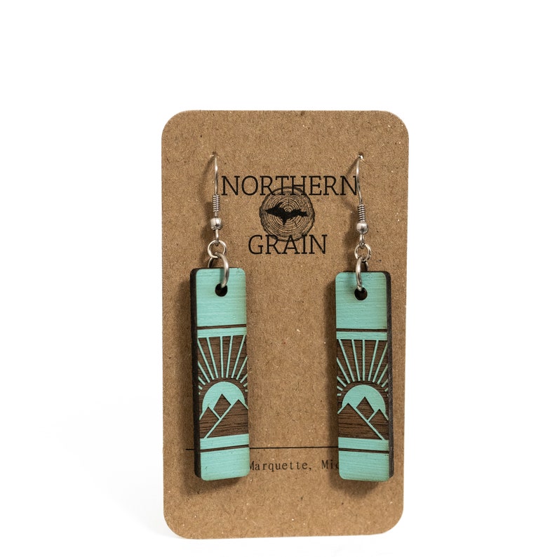 K&ouml;nnte beinhalten: Rechteckige Ohrringe mit einem Berg- und Sonnendesign. Die Ohrringe sind t&uuml;rkis mit einem dunkelbraunen Design und silbernen Haken. Sie werden auf einer braunen Karte mit dem Text "NORTHERN GRAIN" und einem Logo pr&auml;sentiert.