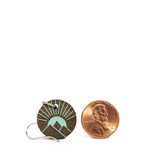 K&ouml;nnte beinhalten: Runder Holzohrring mit Berg- und Sonnendesign in T&uuml;rkis und Braun. Der Ohrring befindet sich neben einem Penny zum Gr&ouml;&szlig;envergleich. Der Ohrring hat einen silbernen Haken.