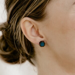 Peut inclure: Gros plan sur une boucle d'oreille en bois avec un cadre octogonal marron fonc&eacute;. Le centre de la boucle d'oreille pr&eacute;sente un motif de couleur turquoise. La boucle d'oreille est port&eacute;e &agrave; l'oreille d'une personne.