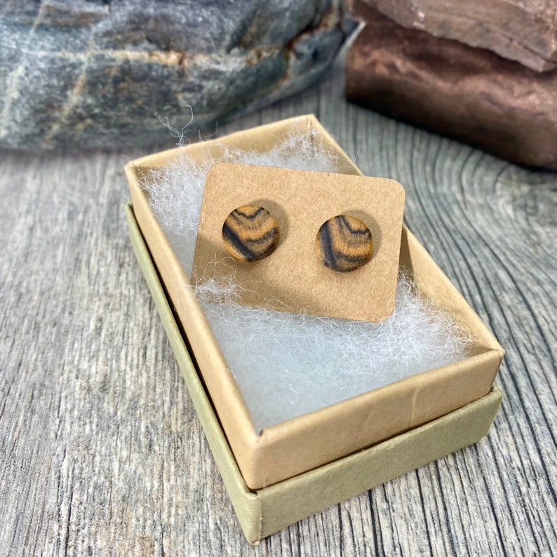 Pu&ograve; includere: Un paio di orecchini rotondi in legno con un motivo a strisce marroni e nere, presentati in una piccola scatola di cartone con carta velina bianca.
