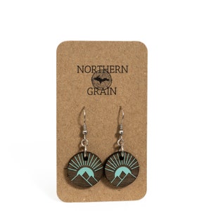 K&ouml;nnte beinhalten: Runde Holzohrringe mit einem t&uuml;rkisfarbenen Berg- und Sonnendesign, die an silberfarbenen Haken h&auml;ngen. Die Ohrringe werden auf einer braunen Karte mit dem Text "NORTHERN GRAIN" pr&auml;sentiert.
