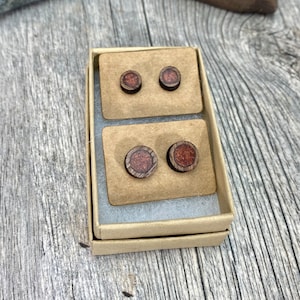 May include: A set of four wooden earrings in a small cardboard box. The earrings are round and have a brown wood base with a red and brown wood inlay.