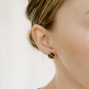 Pu&ograve; includere: Un paio di piccoli orecchini rotondi in legno con un motivo a righe. Gli orecchini sono indossati nei lobi delle orecchie di una persona con i capelli castano chiaro.