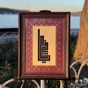 Op de afbeelding: Ingelijst kunstwerk met zwarte kalligrafie op een beige achtergrond, omlijst door een bordeauxrode en gouden patroon. De lijst is donkerbruin. Het kunstwerk wordt tentoongesteld op een zwart schildersezel, met een schilderachtige achtergrond van water en lucht.