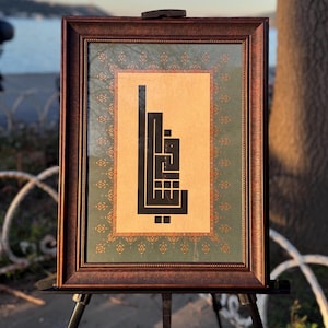 Op de afbeelding: Ingelijst kunstwerk met zwarte kalligrafie op een beige achtergrond, omlijst door een decoratieve groene en rode rand. Het kunstwerk zit in een donkerbruine houten lijst en wordt tentoongesteld op een zwart schildersezel. De achtergrond toont een waterpartij en een boom.