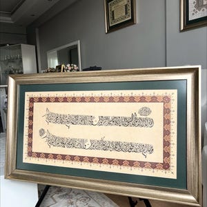 May include: A framed artwork featuring ornate calligraphy in black ink on a beige background, framed with a gold and green border. The artwork is displayed on a black easel, with two other framed pieces visible in the background.