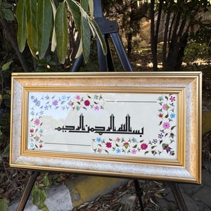 Op de afbeelding: Een ingelijst kunstwerk met een gouden rand, met Arabische kalligrafie in zwart schrift. Het kunstwerk is omgeven door een bloemenrand met roze, blauwe en paarse bloemen. Het frame wordt op een zwart schildersezel weergegeven.