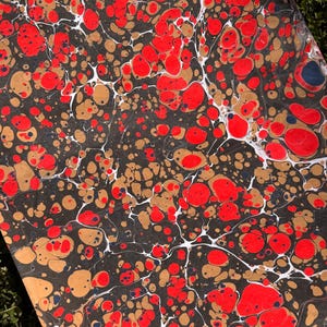 May include: Close-up of a marbled paper sheet with a dark background. The design features red, gold, and white organic shapes. The red shapes are the most prominent, with gold and white accents creating a unique pattern.