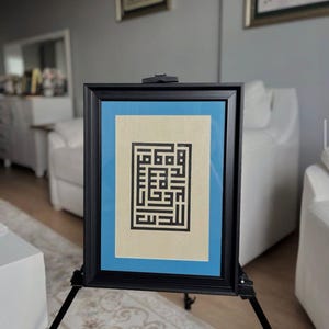 Op de afbeelding: Een ingelijst kunstwerk met zwarte geometrische kalligrafie op een crèmekleurige achtergrond, geplaatst tegen een lichtblauwe passe-partout. De zwarte lijst wordt gepresenteerd op een zwart schildersezel, geschikt voor interieurdecoratie.