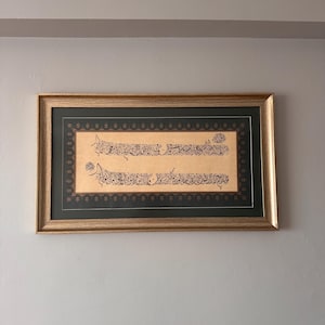 May include: A framed piece of calligraphy art. The artwork features elegant black script on a tan background, set within a dark green mat and a gold-colored frame. The piece is mounted on a light gray wall.