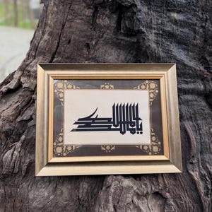 Op de afbeelding: Een ingelijst kunstwerk met zwarte Arabische kalligrafie op een beige achtergrond. De gouden lijst heeft een decoratieve rand. Het kunstwerk wordt gepresenteerd tegen een getextureerde, donkerbruine boomstam.
