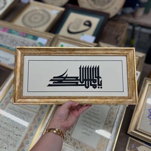 Op de afbeelding: Een ingelijst kunstwerk met zwarte kalligrafie op een crèmekleurige achtergrond. De lijst is goudkleurig en rechthoekig. Op de achtergrond zijn andere ingelijste kunstwerken met vergelijkbare ontwerpen te zien, wat wijst op een verzameling islamitische kunst.